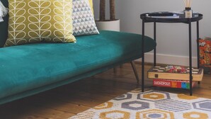 Un salón con un sofá color turquesa, cojines estampados, una planta verde grande, una mesita auxiliar negra con un juego de Monopoly y una alfombra con diseño.