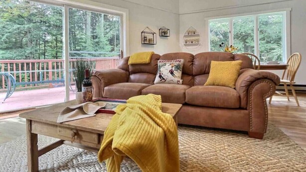 A living room with a brown leather sofa, a wooden coffee table, and a yellow throw blanket. There are two windows with a view of trees and a balcony. - BearVaria AC, Hot Tub, Fire Pit Grill, FrisbeeGolf (Killington)
