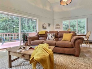 Un salon avec un canapé en cuir marron, une table basse en bois et un plaid jaune. Il y a deux fenêtres donnant sur des arbres et un balcon.