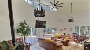 A spacious living room with a fireplace, leather sofas, and a large window overlooking a deck. - BearVaria AC, Hot Tub, Fire Pit Grill, FrisbeeGolf (Killington)