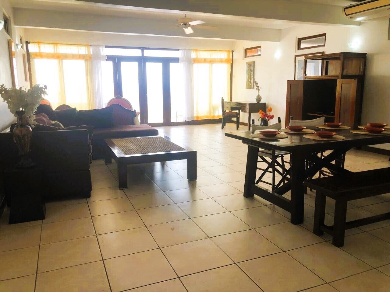 A living room with a sofa, coffee table, and dining table with chairs. There is a large window with curtains, a ceiling fan, and a television on a wooden cabinet.