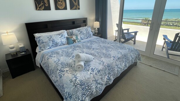Habitación de hotel con una cama grande, una mesita de noche con lámpara y vista al océano a través de un ventanal corredero.