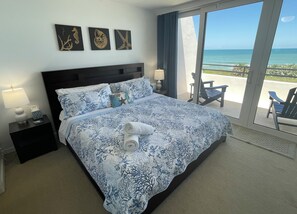 A hotel room with a large bed, a nightstand with a lamp, and a view of the ocean through a sliding door. - Flagler Beach, Florida<br>Ocean-View Townhome<br> Aliki Sunrise Surprise! 🚀🌊🏖️☀️ (Flagler Beach)