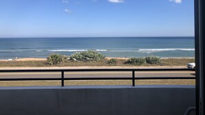 Vista del océano desde un balcón con barandilla, cielo despejado y una playa distante.