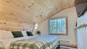 Une chambre avec un plafond en bois, un lit recouvert d’une couverture à carreaux, une fenêtre donnant sur les arbres et une étagère intégrée.
