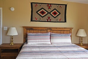 Een slaapkamer met een houten hoofdbord, een gestreept dekbedovertrek, twee nachtlampjes en een wandtapijt.