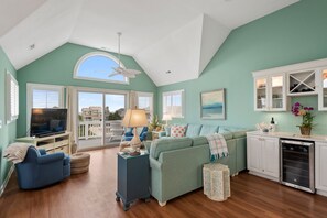 A spacious living room with a large TV, comfortable seating, and a kitchen area with cabinets and appliances. - Welcome to MAJESTY! Ocean Views-Hot Tub-Pool Access (Nags Head)
