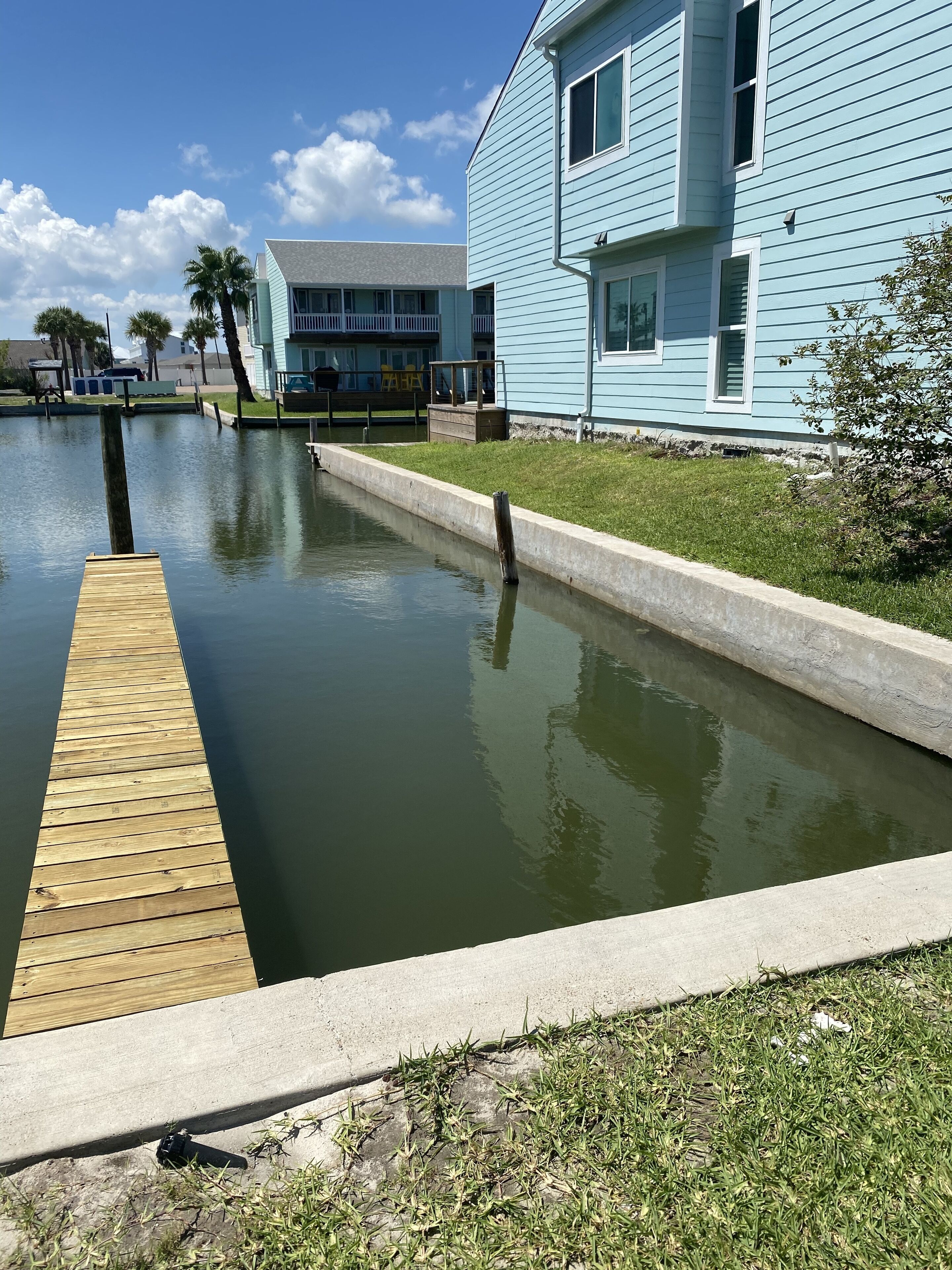 A waterfront property with a wooden dock extending into a calm body of water, surrounded by a grassy area and a light blue building with multiple windows.