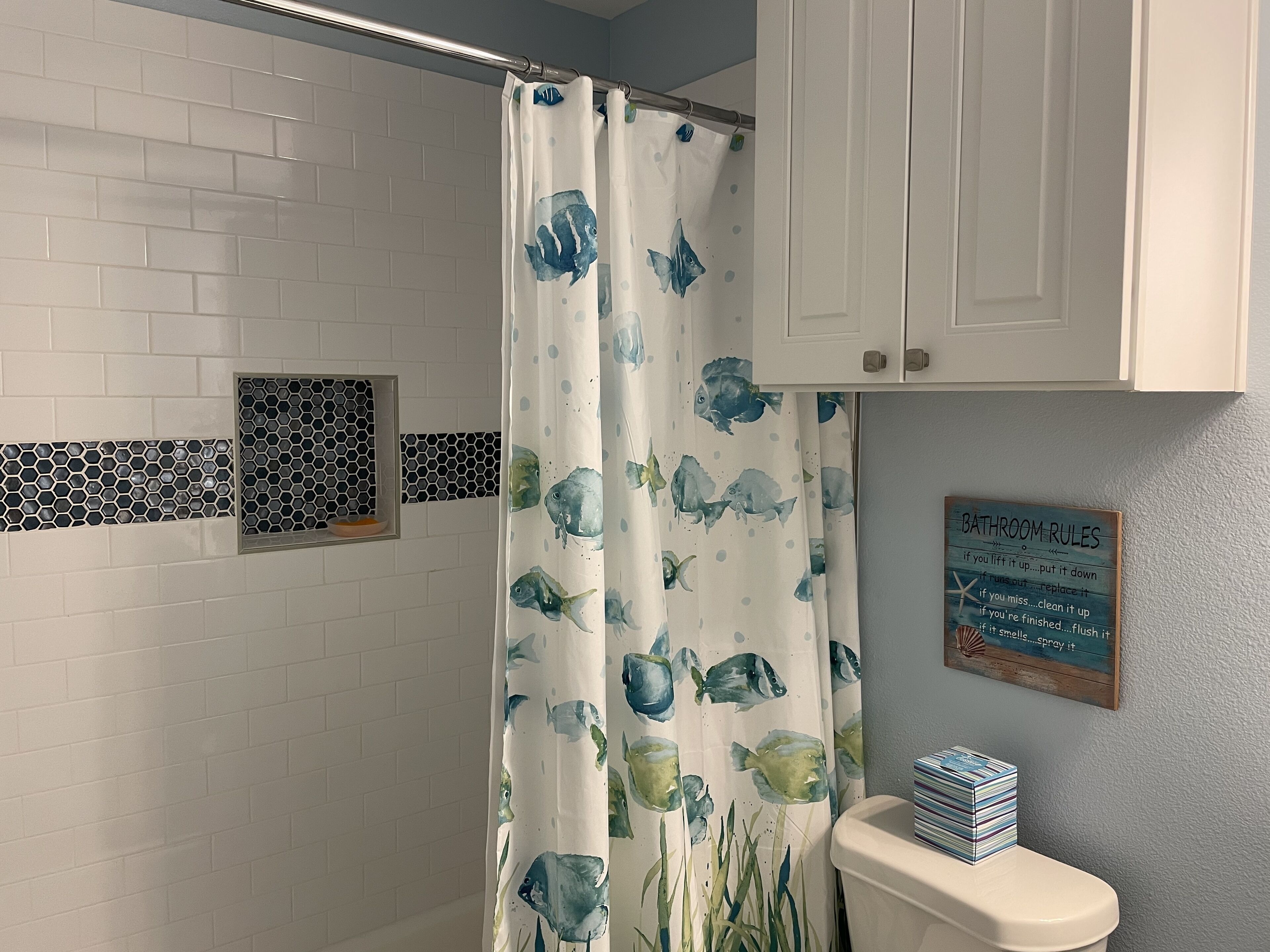 A bathroom with a shower curtain featuring fish patterns, a toilet, a small shelf with a bowl, and a sign with bathroom rules.