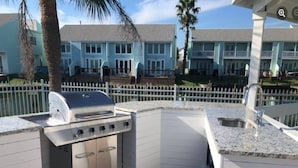 An outdoor kitchen with a grill, sink, and storage cabinets, surrounded by a white fence and palm trees.