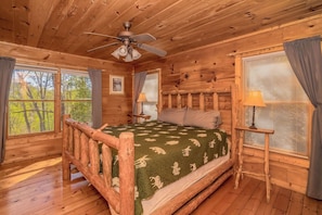 A log cabin bedroom with a bed, a ceiling fan, and windows with curtains.