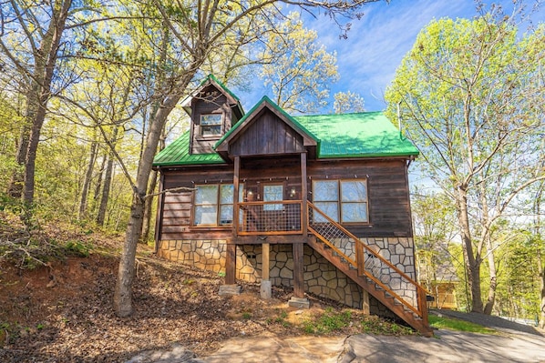 A cabin with a stone foundation, wooden exterior, and a green roof, surrounded by trees.