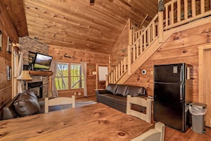 A wooden cabin interior with a dining area, a sofa, a television, and a staircase.