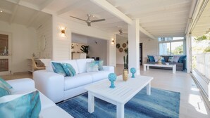 Un'ampia zona giorno con un divano bianco, un tavolino da caffè e un tappeto blu. È presente una ventola a soffitto e una grande finestra che offre luce naturale.