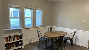 A room with a wooden floor, a round table with four chairs, a bookshelf, and three windows with blinds. - Perfect Location Just 0.5 Miles from Notre Dame’s Campus! (South Bend)