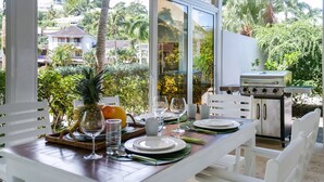 An outdoor dining area with a view of palm trees and buildings, a grill, and a table set with plates, glasses, and fruits. - AVRAMAR VILLA PLAYA LAS BALLENAS LAS TERRENAS (Las Terrenas)