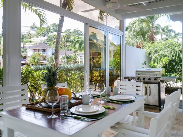 An outdoor dining area with a view of palm trees and buildings, a grill, and a table set with plates, glasses, and fruits.