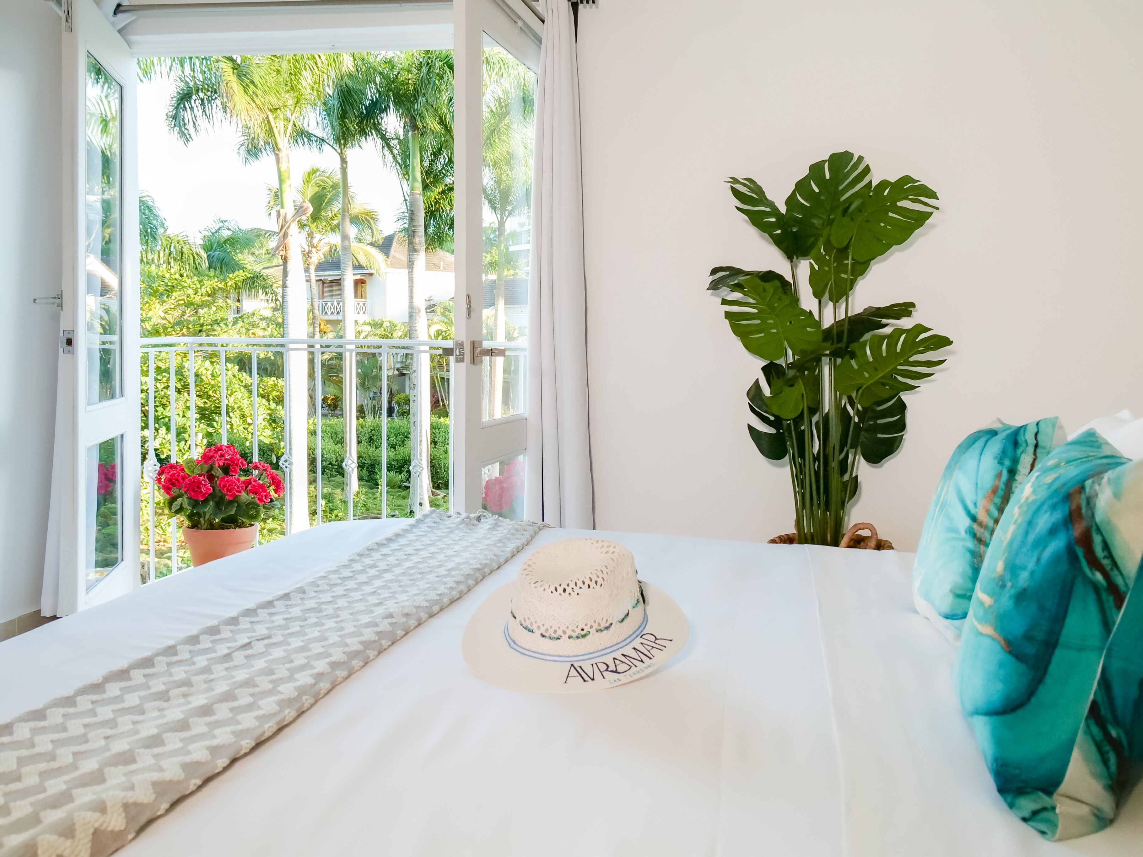 Varanda com vista para palmeiras e um jardim, um vaso de planta e uma cama com chapéu e manta azul.