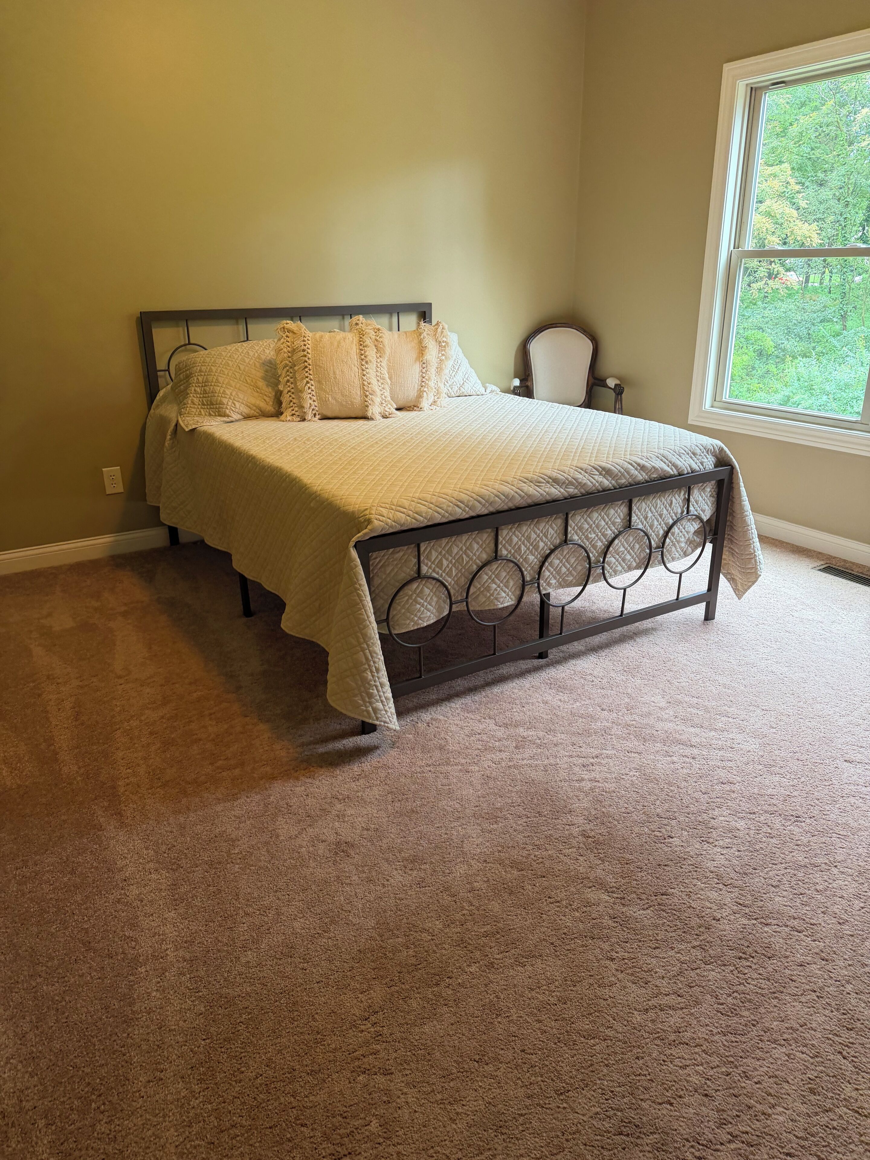 A bedroom with a bed, carpeted floor, and a window with a view of greenery.