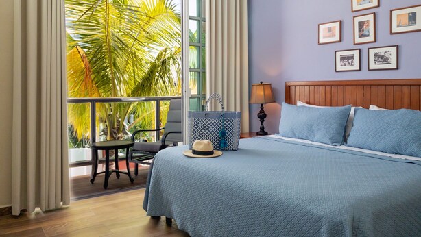 Une chambre d’hôtel avec un grand lit, un balcon agrémenté de palmiers, une table avec une chaise et des tableaux encadrés au mur.
