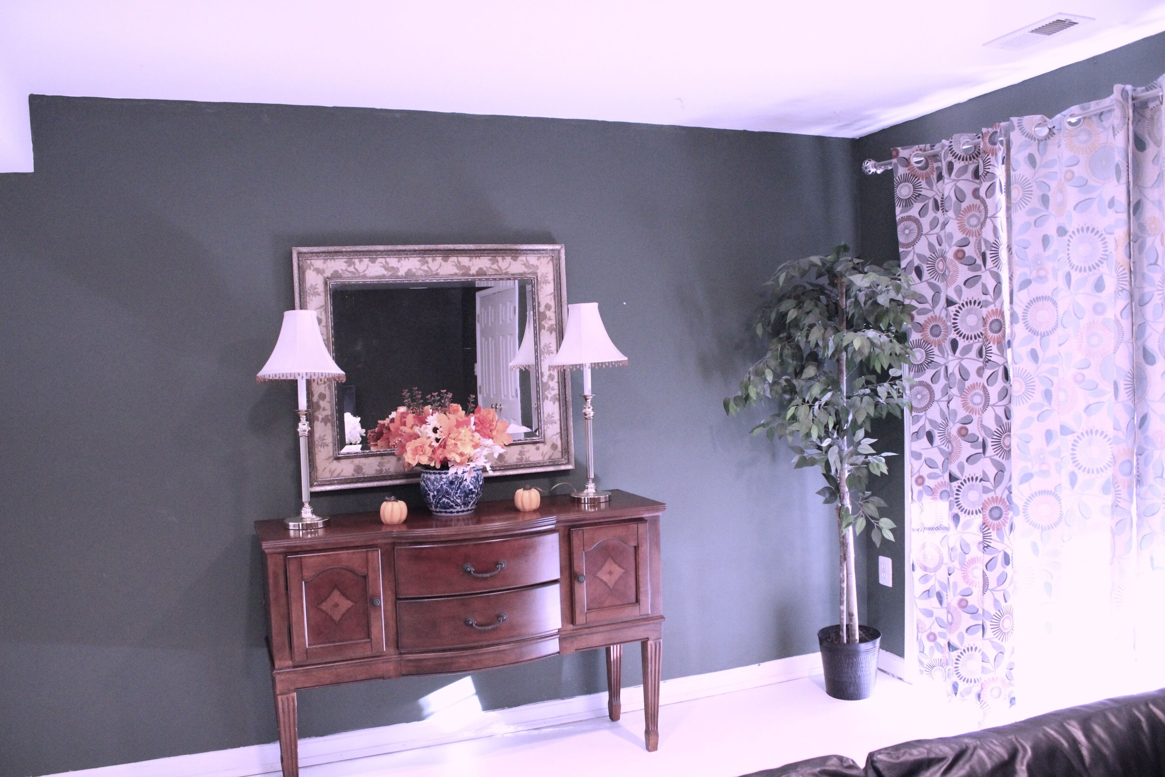 Una habitación con una pared oscura, un aparador de madera con dos lámparas, un espejo y una planta en maceta.