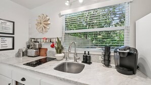 A modern kitchen with a sink, stove, and coffee maker. There is a window with blinds and a decorative wall piece. - Pet Friendly Studio with Private Fenced Yard (Jacksonville)