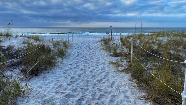Ein sandiger Pfad am Strand, der zum Meer führt, mit einem bewölkten Himmel.