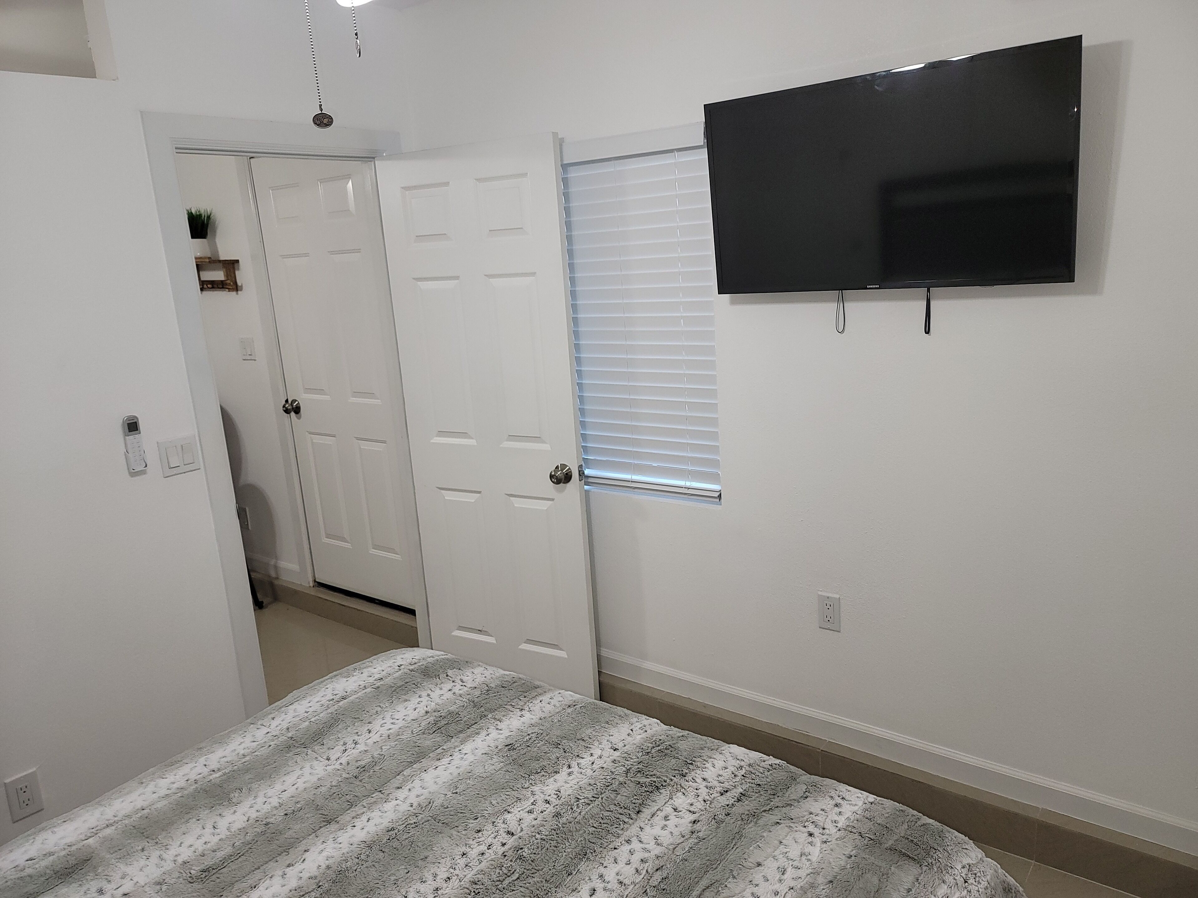 Quarto com cama, televisão fixada na parede, porta e janela com persianas.