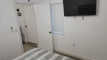 Ein Schlafzimmer mit einem Bett, einem an der Wand montierten Fernseher, einer Tür und einem Fenster mit Jalousien.