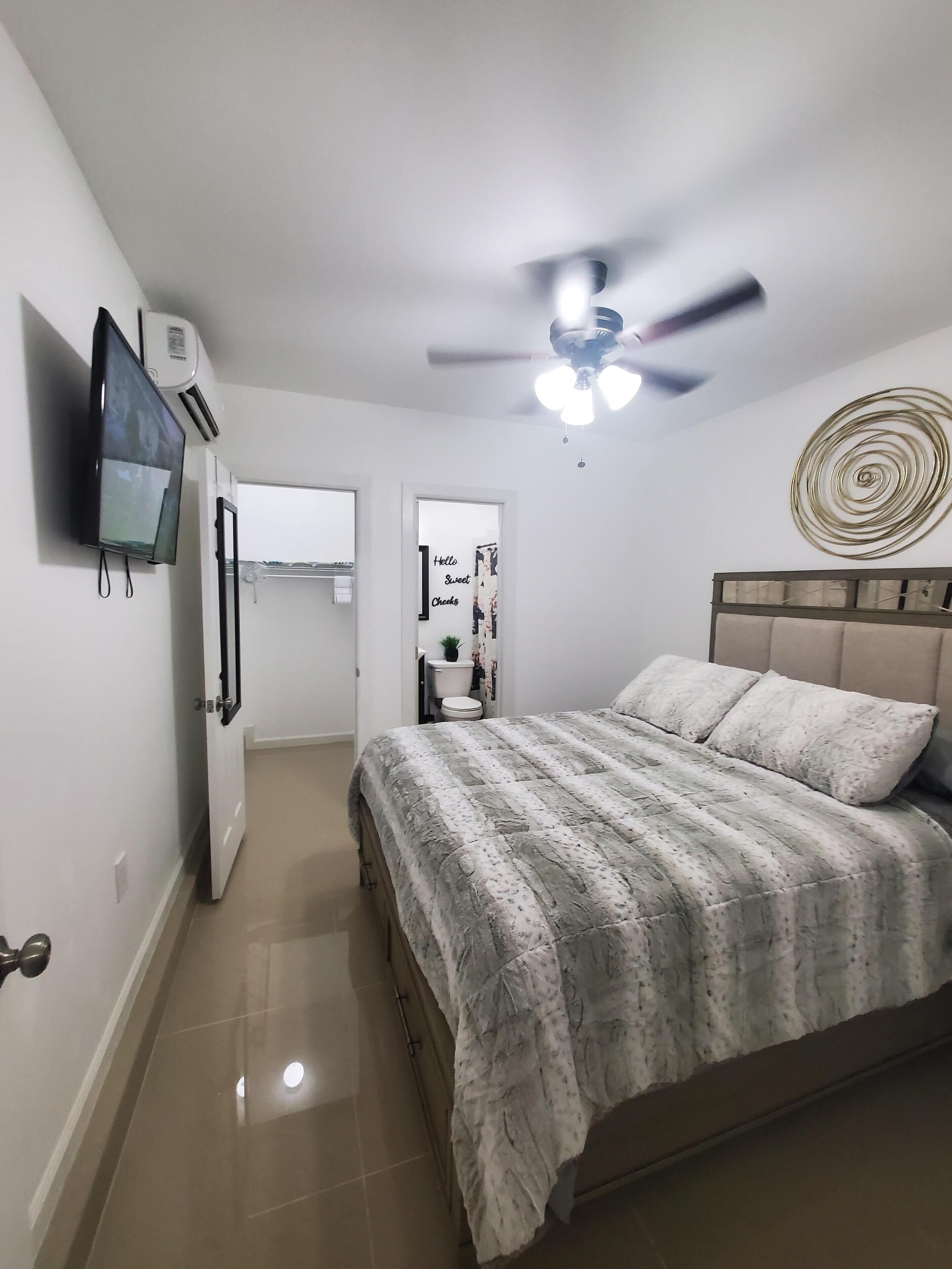 A bedroom with a bed, a ceiling fan, a wall-mounted TV, and a door leading to a bathroom.