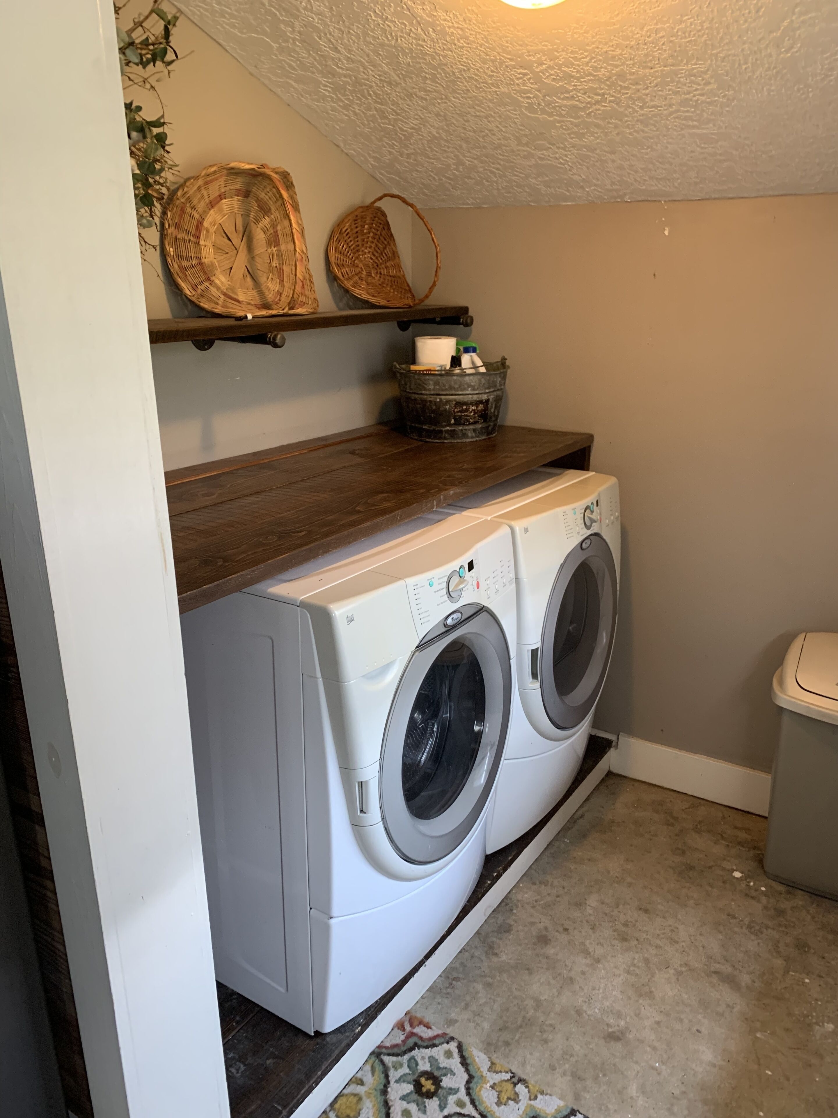 A laundry room with two front-loading washing machines, a wooden shelf with baskets, and a trash can.