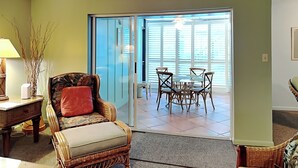 A living room with a wicker sofa, a small table with a lamp, and a dining area with a glass table and chairs. - Jacksonville Beach Costa Verde 2319-102 by Distinctive Beach Rentals (Jacksonville Beach)