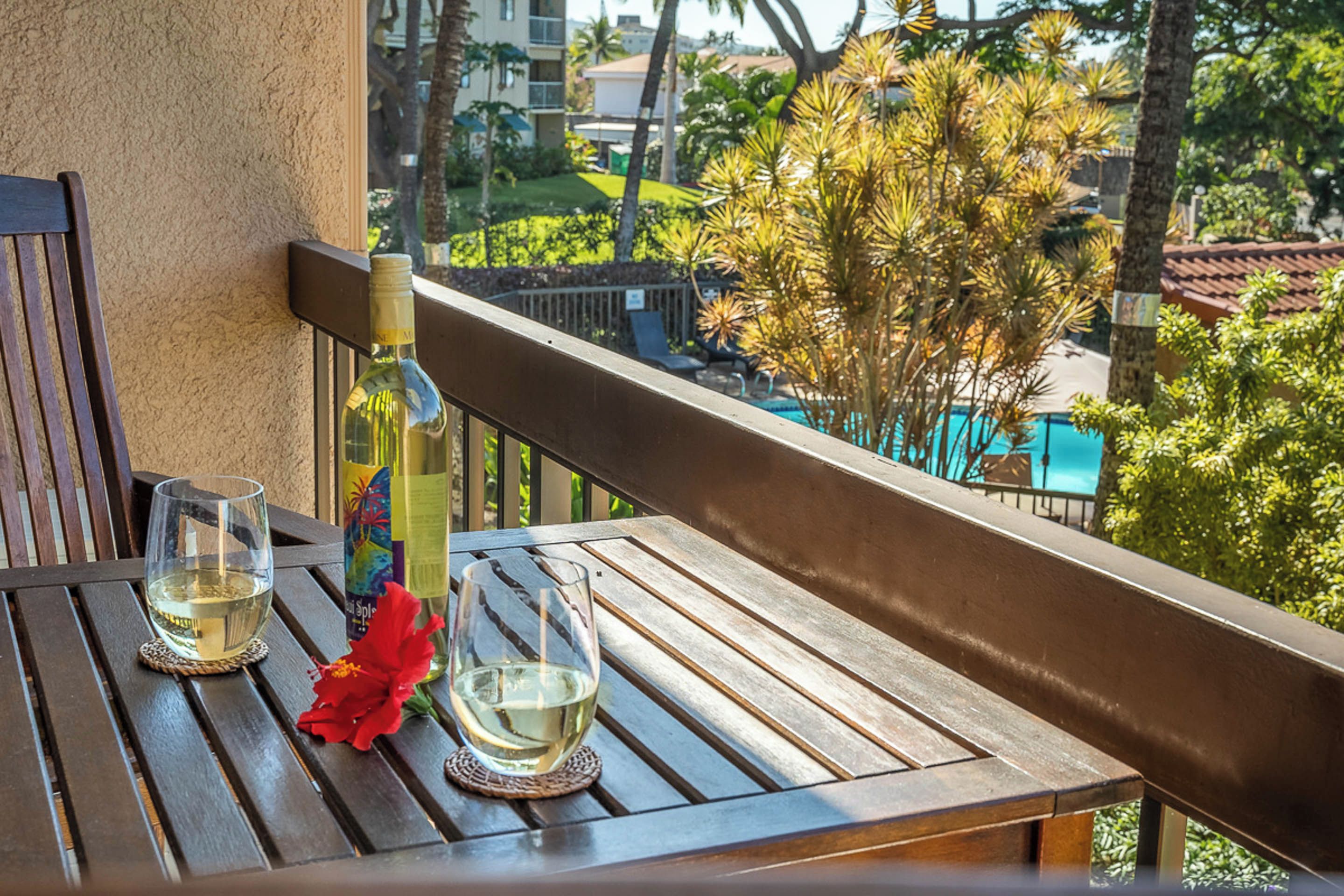 A wooden table with two glasses of white wine and a bottle of wine on a balcony overlooking a pool and greenery.