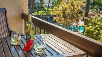 A wooden table with two glasses of white wine and a bottle of wine on a balcony overlooking a pool and greenery.