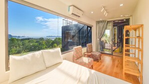 A bedroom with a large window, a bed, two floral chairs, a wooden shelf, and a view of greenery and distant structures. - Tuan Chau Ruby Villa (Tuan Chau)