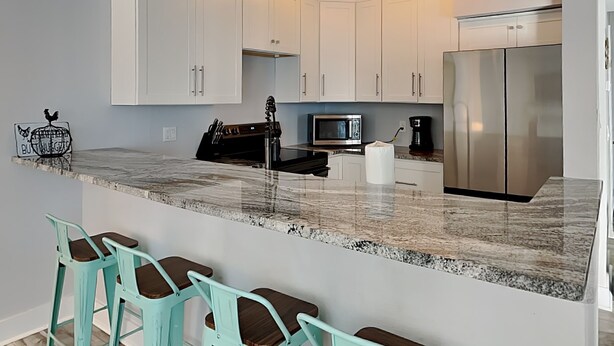 A kitchen with white cabinets, a stainless steel refrigerator, a microwave, and a coffee maker. There are bar stools with turquoise seats. - Madeira Beach Yacht Club 215F by Distinctive Beach Rentals (Madeira Beach)