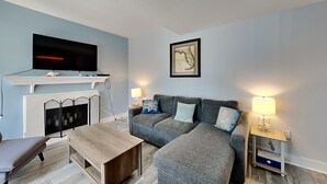 A modern living room with a grey sofa, a wooden coffee table, a flat-screen TV mounted above a fireplace, and two bedside lamps. - Madeira Beach Yacht Club 215F by Distinctive Beach Rentals (Madeira Beach)