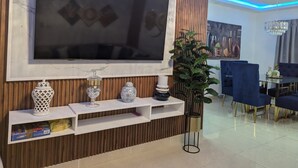 Ein modernes Wohnzimmer mit einem großen Flachbildfernseher an einer Holzwand, einem Essbereich mit blauen Stühlen und einer Kandelaberleuchte.
