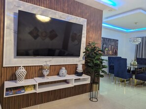 Ein modernes Wohnzimmer mit einem großen Flachbildfernseher an einer Holzwand, einem Essbereich mit blauen Stühlen und einer Kandelaberleuchte.
