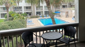 Un balcón con dos sillas y una mesa, con vista a una zona de piscina con sillones y una palmera.