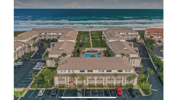 Un hotel frente a la playa con varias edificaciones, estacionamiento y zona de piscina.