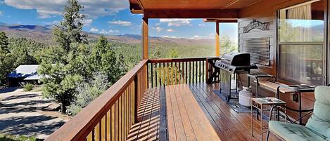 A wooden deck with a view of mountains, a grill, and a table with chairs.