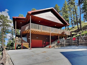 A modern two-story building with a red garage door, a balcony with a wooden railing, and a stone retaining wall. - Ain't No Better View by Ruidoso Vacation Rentals (Ruidoso)