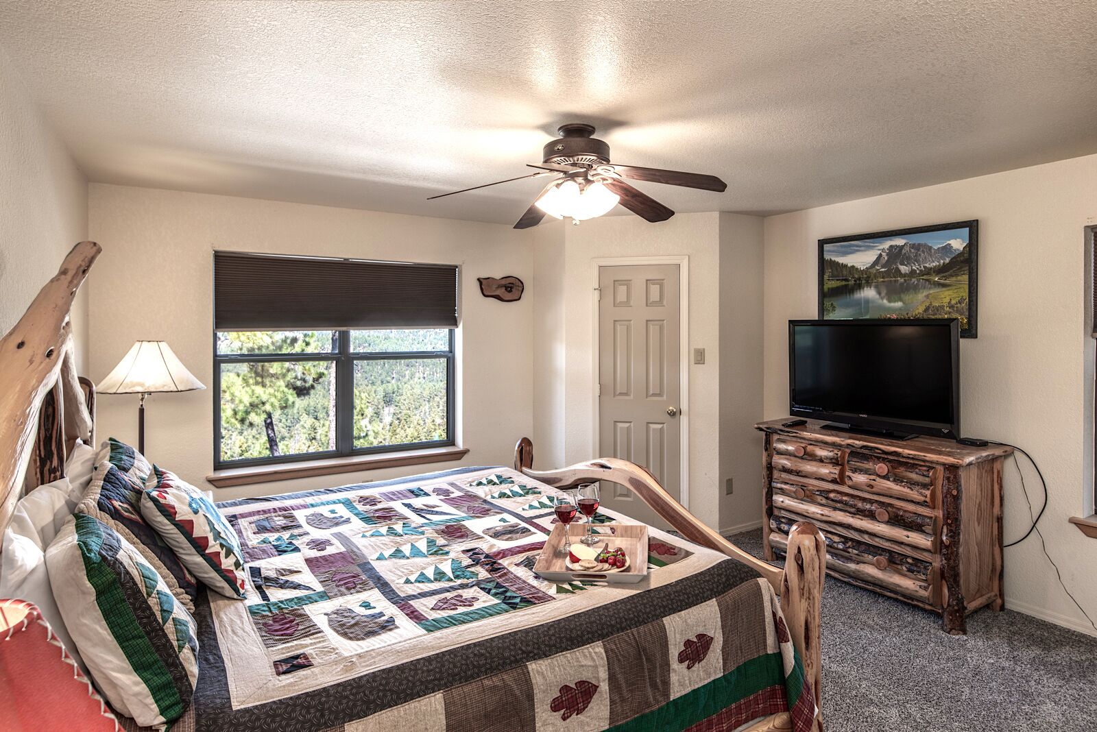 A bedroom with a bed, a ceiling fan, a TV, a window with a view of trees, and a wooden dresser.
