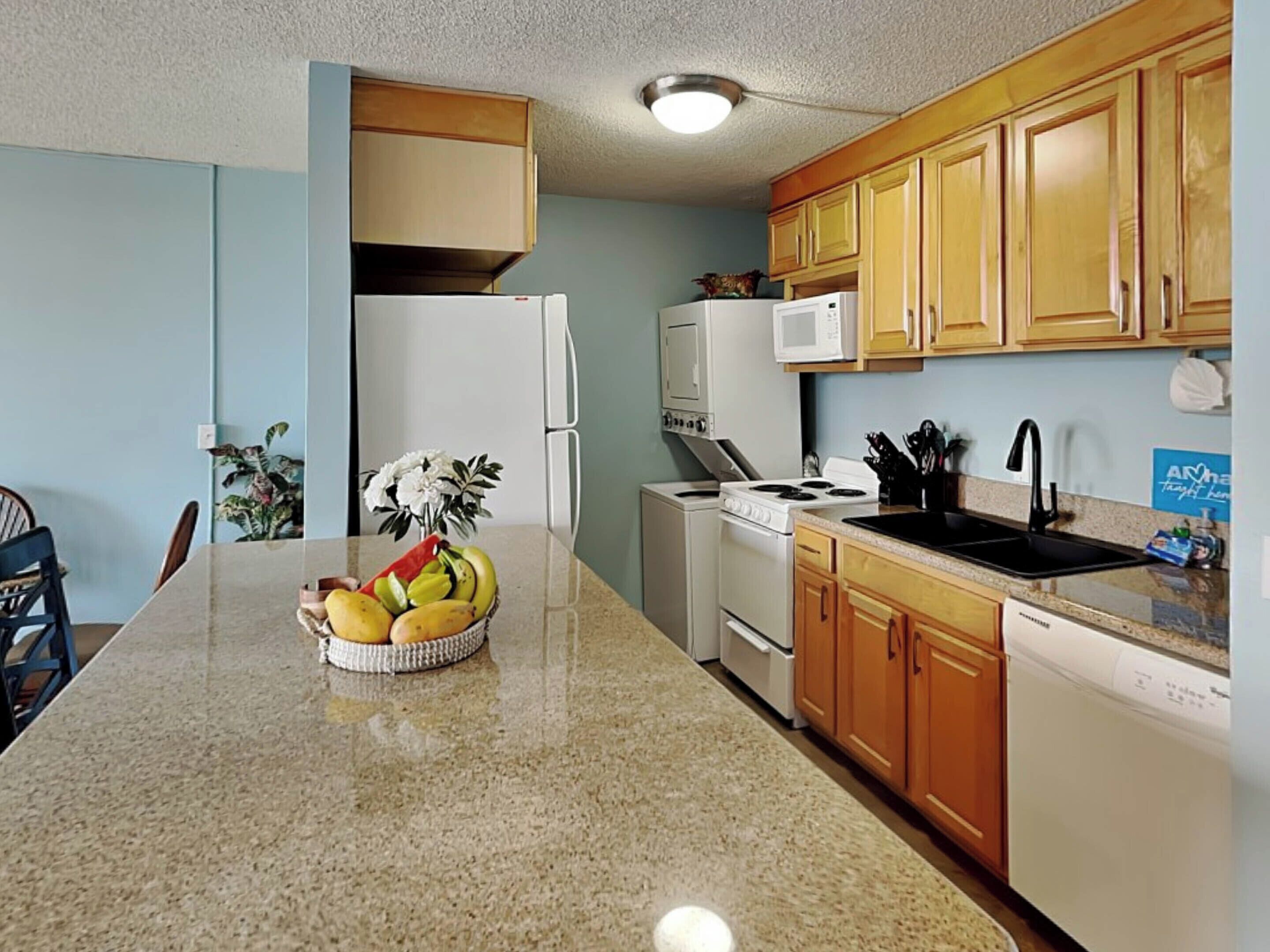A kitchen with a granite countertop, white appliances, wooden cabinets, and a bowl of fruit on the island.