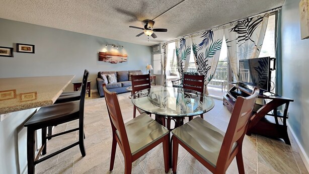 A dining area with a glass table, wooden chairs, a ceiling fan, and a sofa in the background. - Island Surf 312 by Maui Rental Group (Kihei)