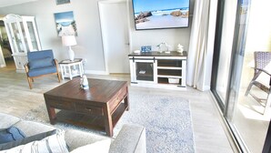 Una sala de estar con televisor, una mesa de centro, un sillón azul, una lámpara y vista a la playa por la ventana.