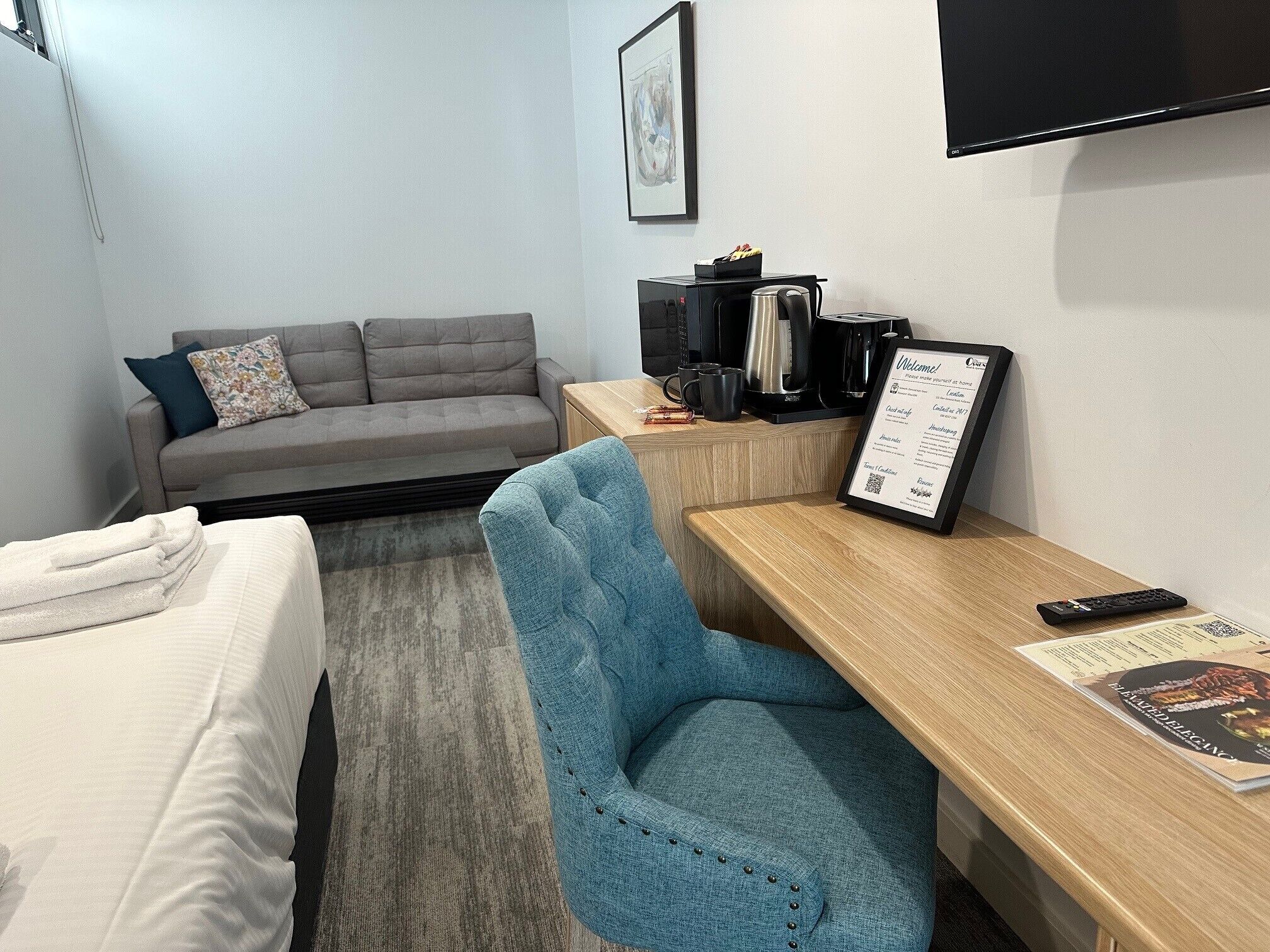 Een hotelkamer met een bed, een blauwe fauteuil, een houten bureau, een koffiezetapparaat en een bord met een welkomstboodschap.