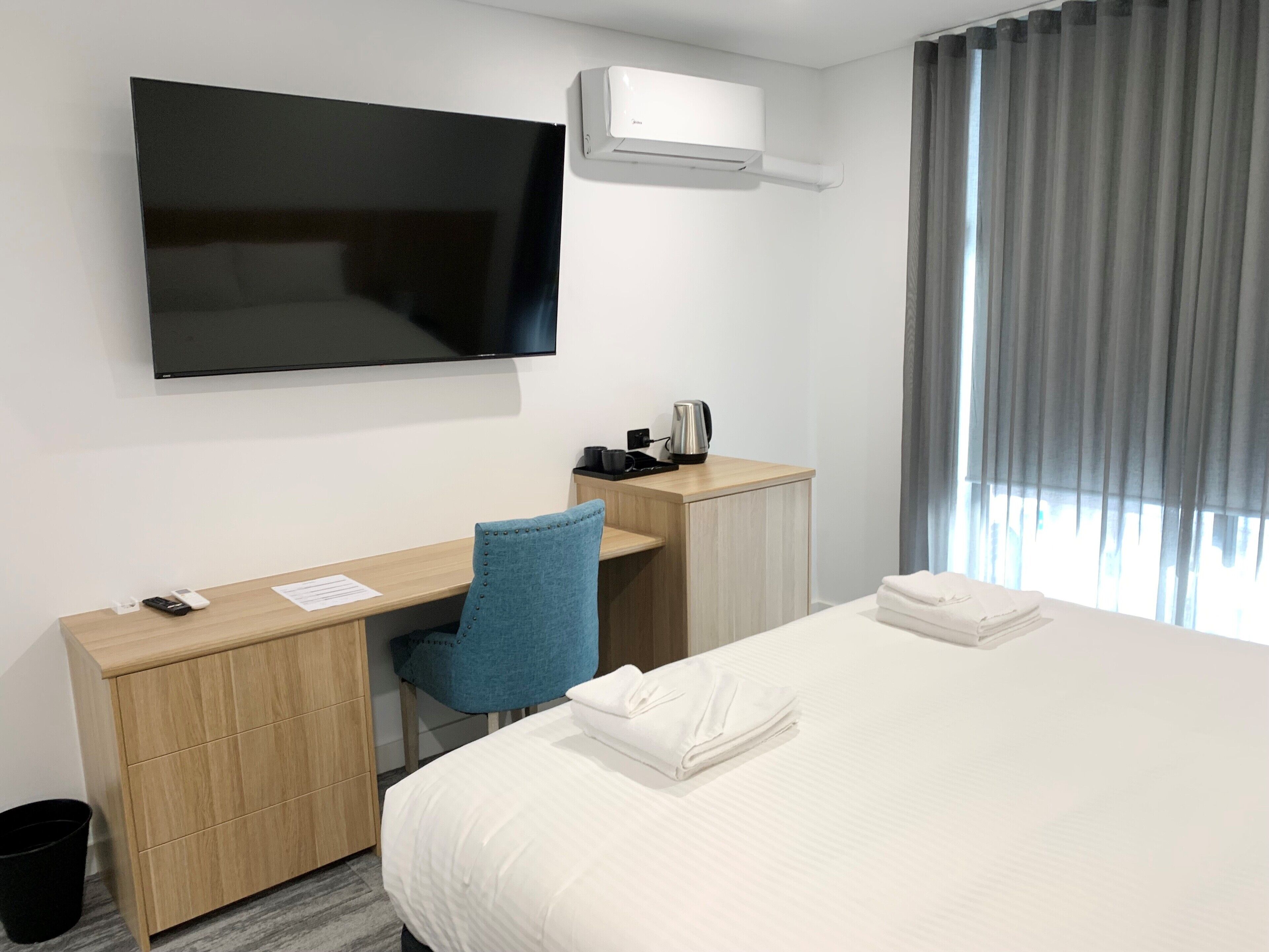A hotel room with a bed, a desk with a chair, a flat-screen TV, a coffee maker, and a window with curtains.