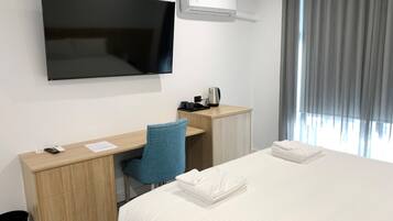A hotel room with a bed, a desk with a chair, a flat-screen TV, a coffee maker, and a window with curtains.